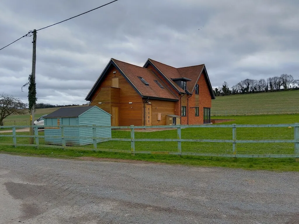 A contemporary timber-clad UK new-build under grey skies with post-and-rail fencing and open countryside beyond — heat pump sizing must match the property's heat load for the system to run at its rated efficiency.